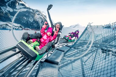 People enjoying a winter sled ride on a track with snow-covered mountains in the background. | © Kitzsteinhorn 