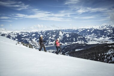 Two people snowshoeing on a snowy mountain slope with a view of mountains and a clear blue sky, heading through the winter landscape. | © Zell am See-Kaprun Tourismus